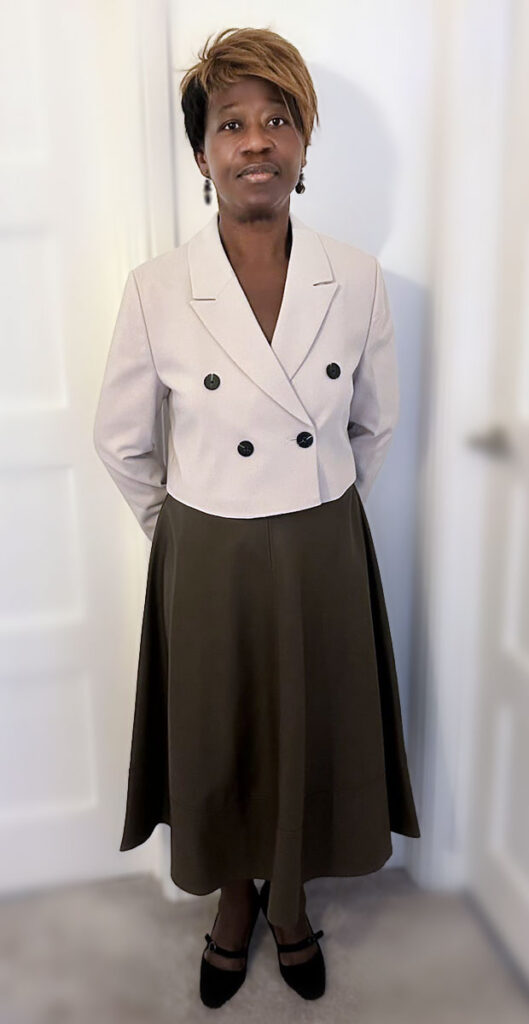 A person stands indoors wearing a formal outfit with a light blazer and dark skirt. The setting is a simple, white-walled room.