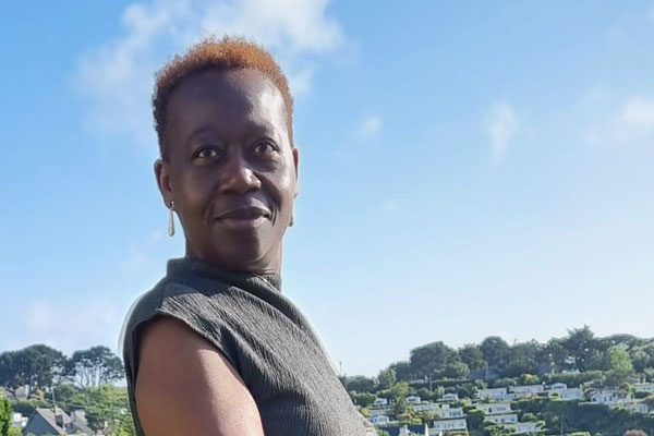 A person stands outdoors with a view of greenery and residential houses in the background under a clear blue sky.