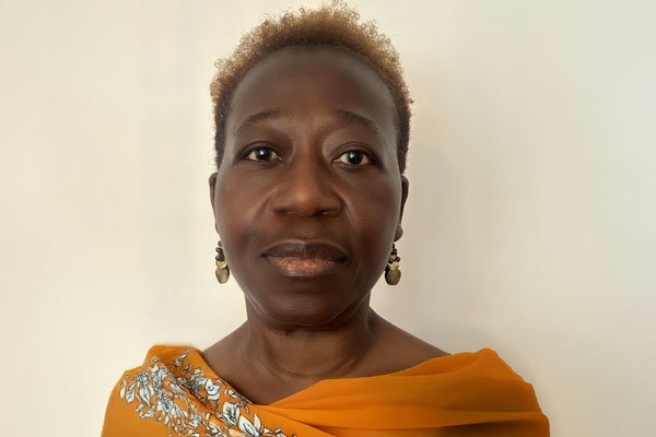 A person wearing an orange and floral-patterned garment stands against a plain background, gazing at the camera, with earrings and short hair.