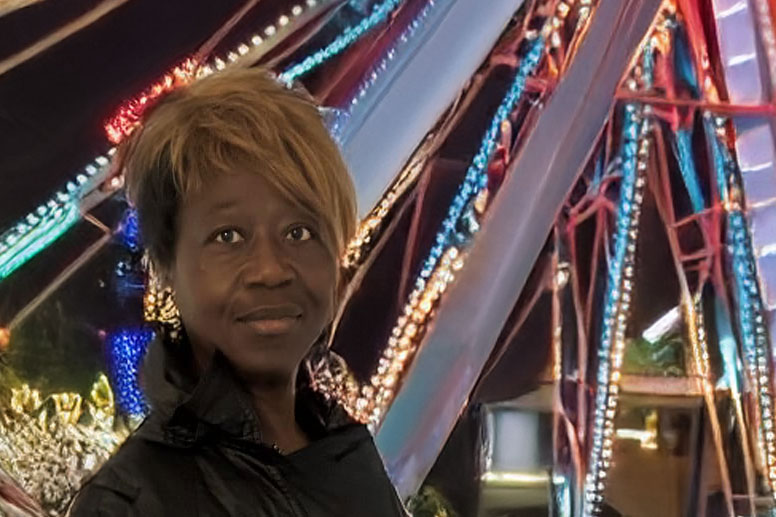 A person with short hair stands near a colorful, illuminated structure, possibly a Ferris wheel, at night. Vibrant lights create a festive atmosphere.