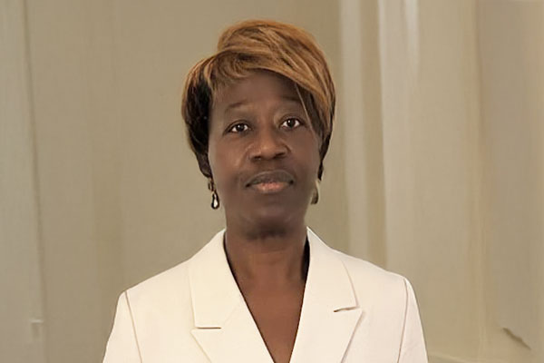 A person in a white outfit stands indoors against a neutral backdrop, looking at the camera. The surroundings appear minimalistic and professional.