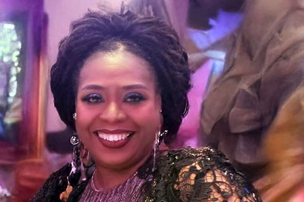 A person smiling indoors, wearing elegant attire with detailed accessories. The background features soft lighting and blurred, artistic decor elements.