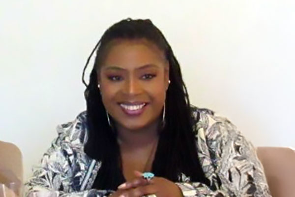 A person with long hair and a patterned jacket smiles while sitting against a plain background. No landmarks or historical buildings are visible.