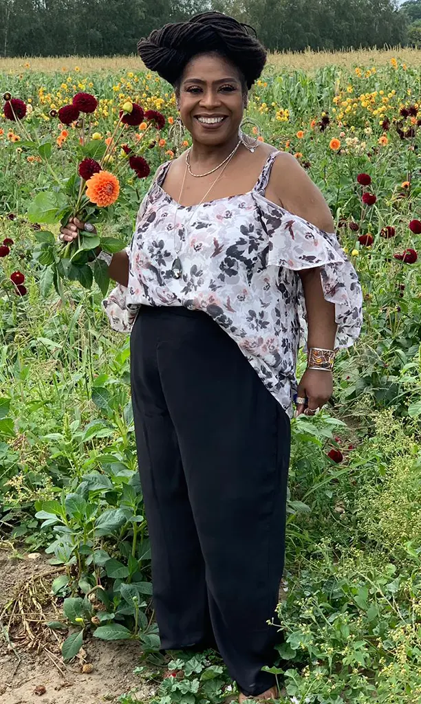 A person stands in a vibrant flower field holding colorful blossoms, wearing a floral top and dark pants, with trees in the background.