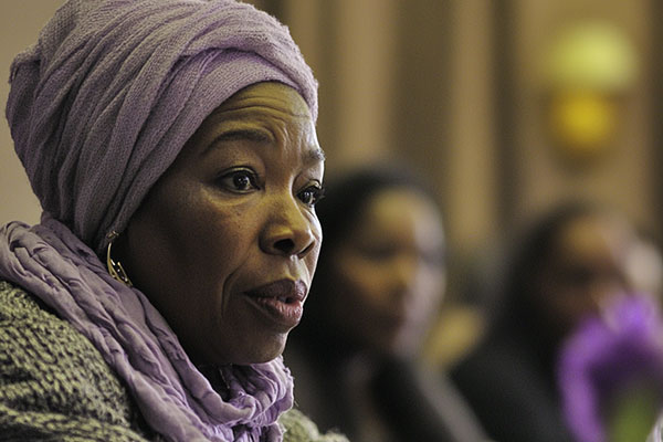 A person wearing a purple headscarf participates in a meeting or discussion, with several other blurred individuals visible in the background.