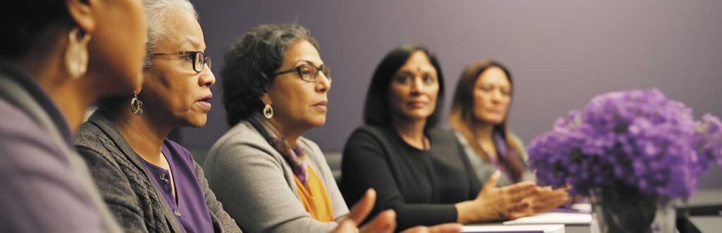 Five people are seated in a meeting room, engaged in discussion. A bouquet of purple flowers is in the foreground.